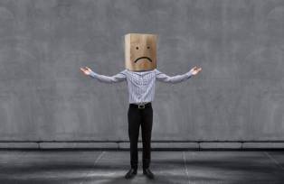Homem com saco de papelão na cabeça, que contém um emoji triste.