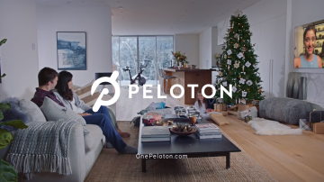 Comercial da Peloton mostra um casal sentado no sofá assitindo televisão. O logo da Peloton aparece centralizado na tela