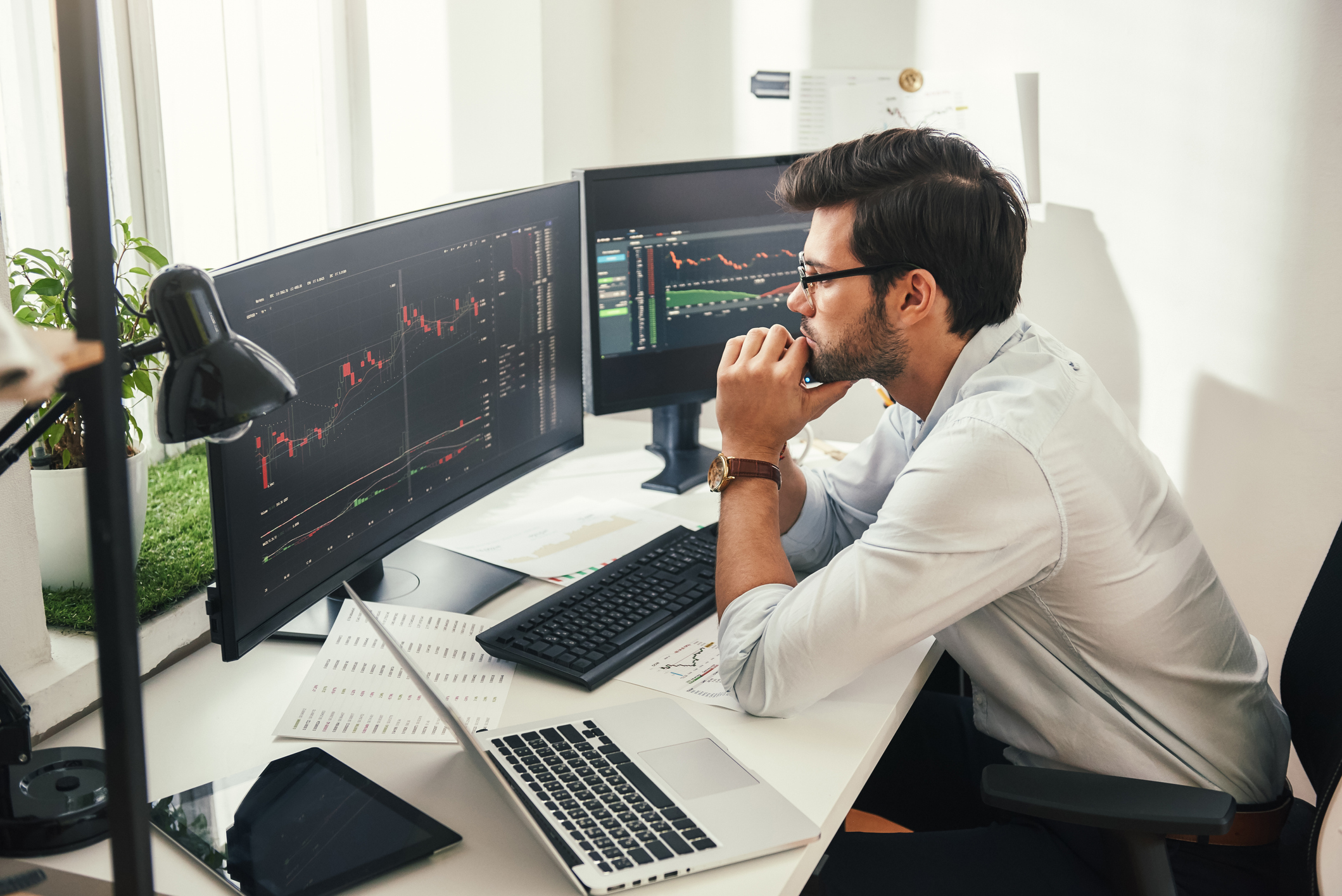 Successful traders. Back view of bearded stock market broker in eyeglasses analyzing data and graphs on multiple computer screens while sitting in modern office. Master Trader