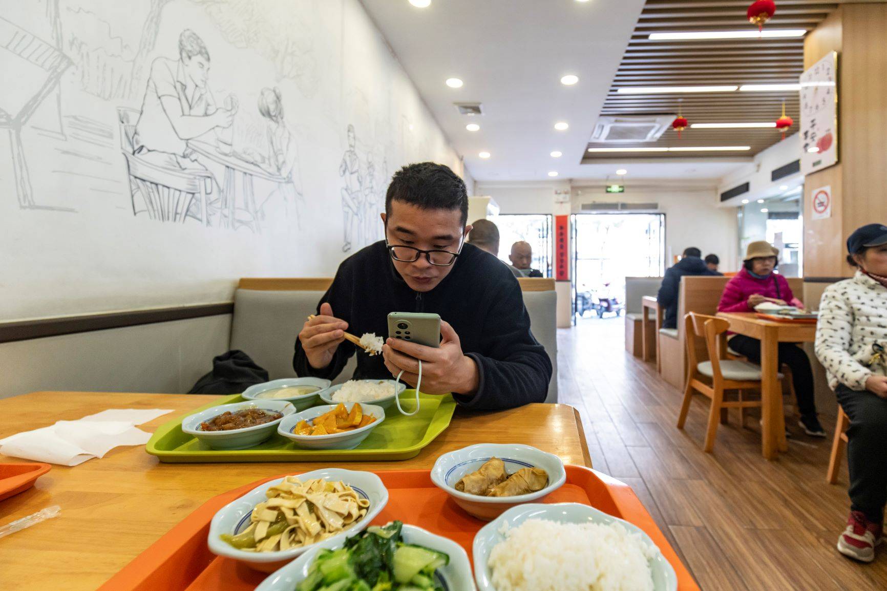 Um jovem profissional olha para seu telefone enquanto come em uma cantina comunitária em Xangai, em 22 de março de 2024. As cantinas comunitárias que oferecem pratos enormes por um ou dois dólares se tornaram populares entre jovens profissionais econômicos. (Qilai Shen/The New York Times)