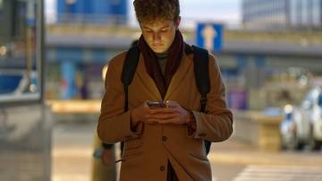 Homem branco com casaco marrom andando com o celular na mão e olhando para a tela